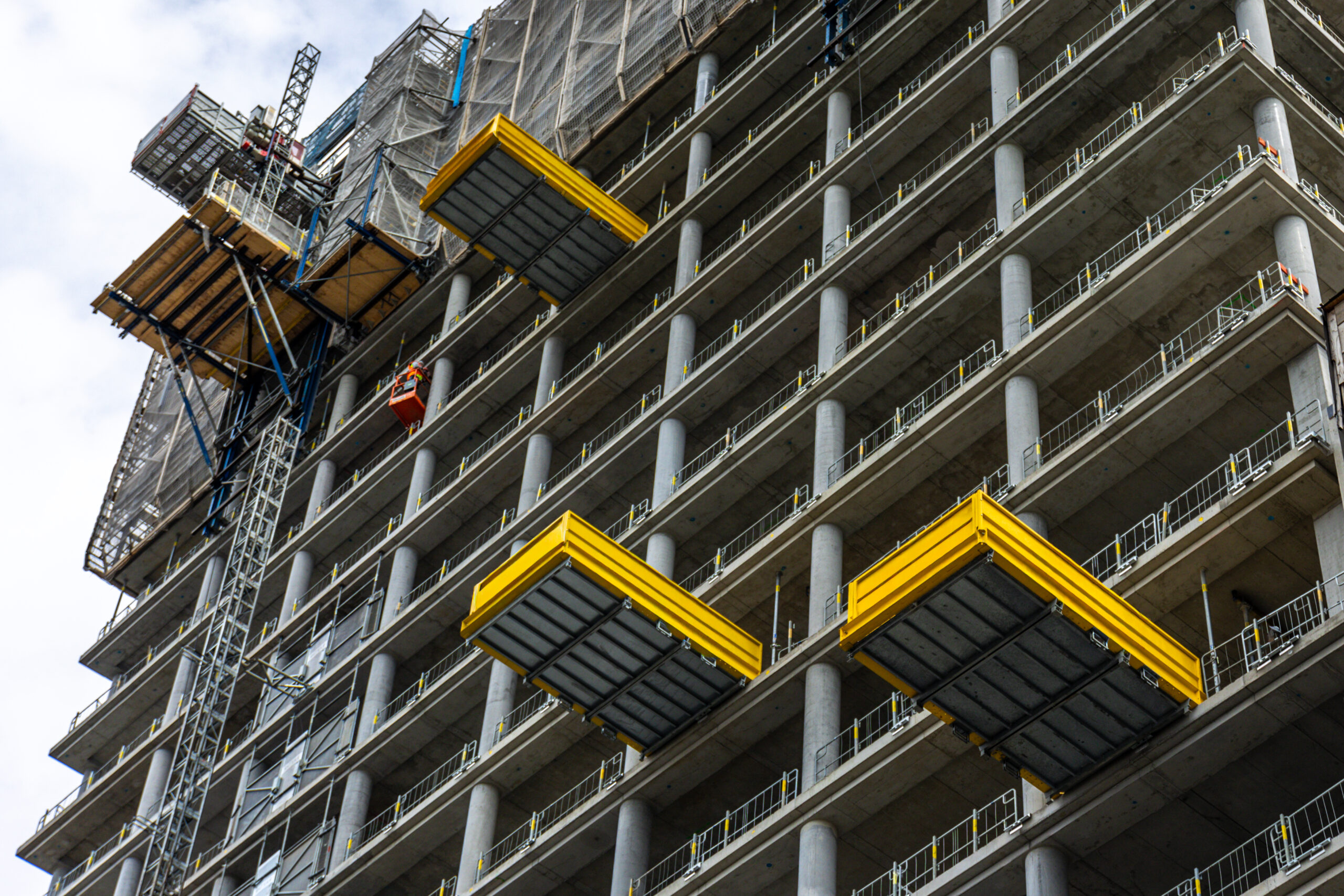 Close view of a high rise section with three yellow cantilever decks, scaffolding and a hoist. Repeating balconies and technical details show modern construction progress.